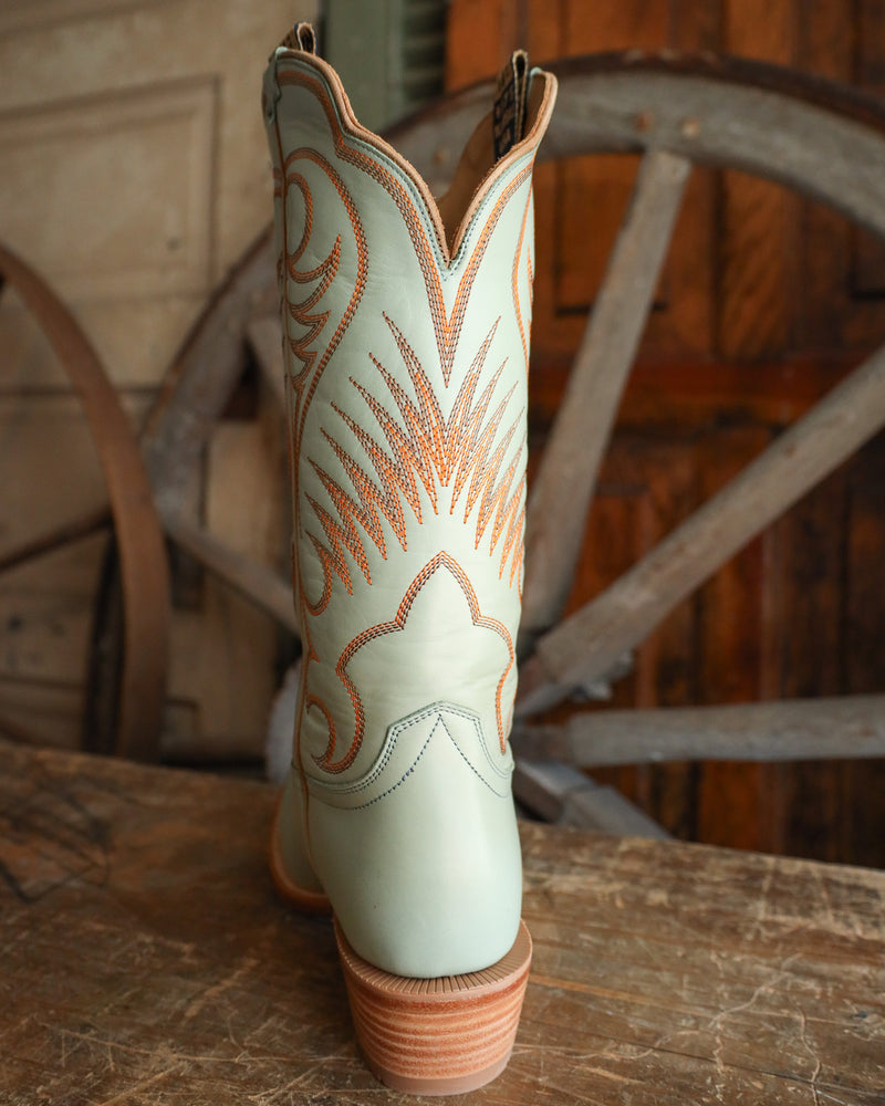 Light green cowboy boot with brown stitching on a wooden surface with a rustic background