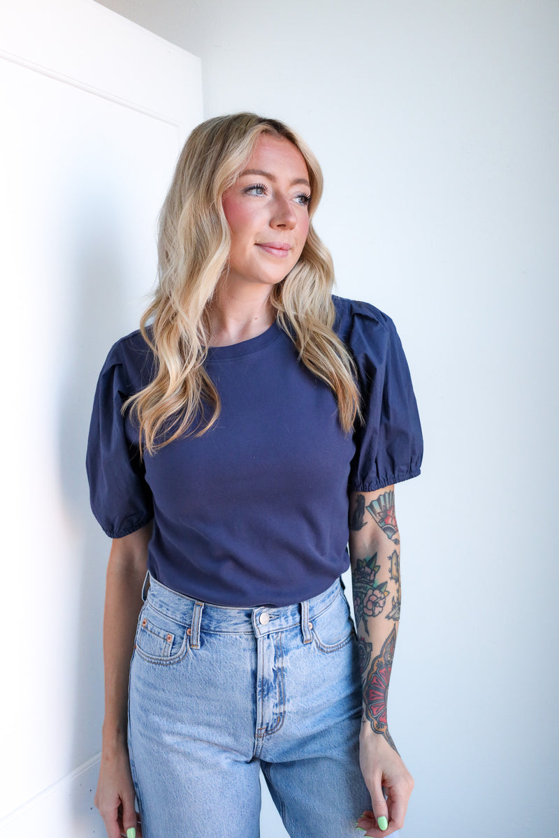 Woman wearing a navy blue top and light blue jeans against a white wall