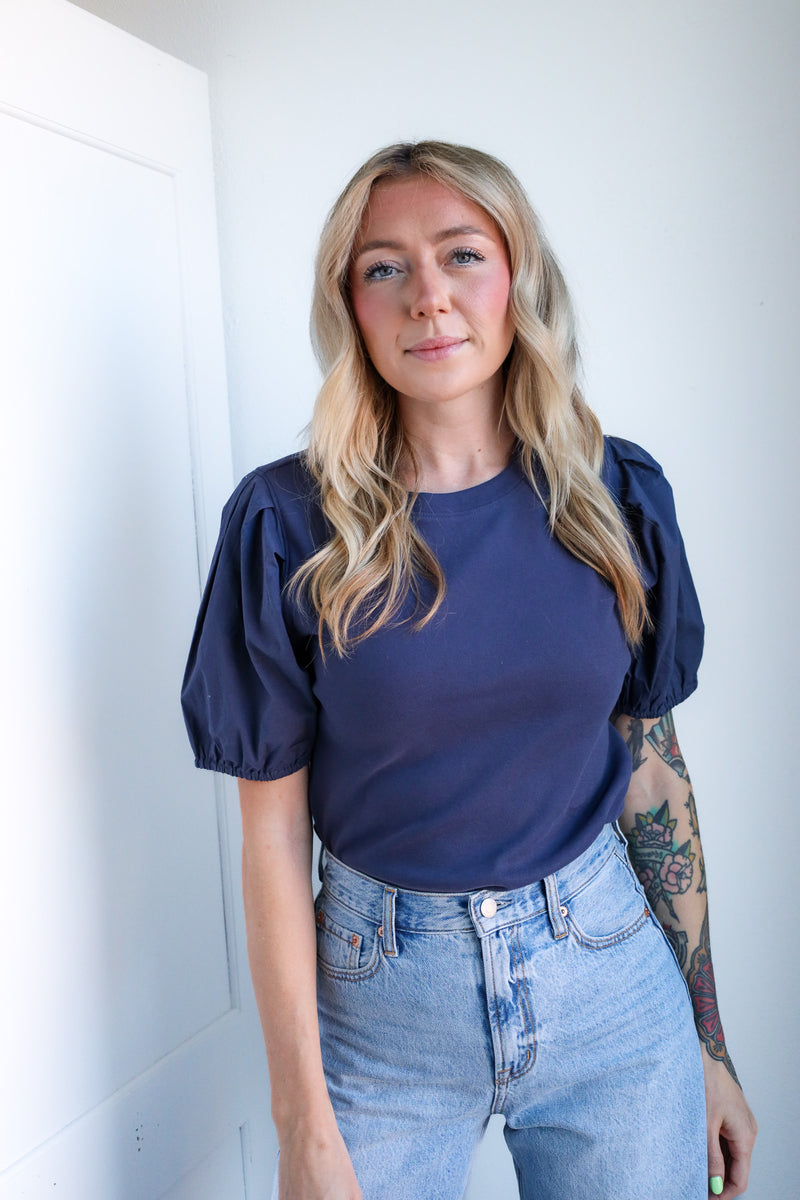 Woman wearing a navy blue top with puffed sleeves and light blue jeans against a white wall.