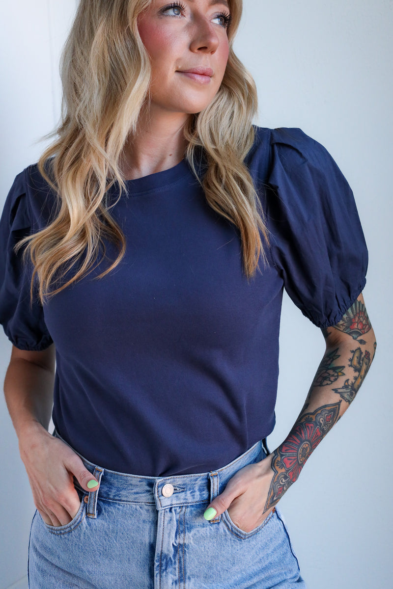 Woman wearing a navy blue top with puffed sleeves and high-waisted jeans against a plain background