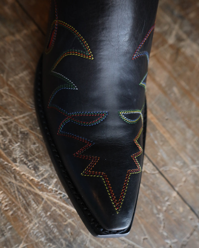 Close-up of a black leather boot with colorful stitching on a wooden floor.