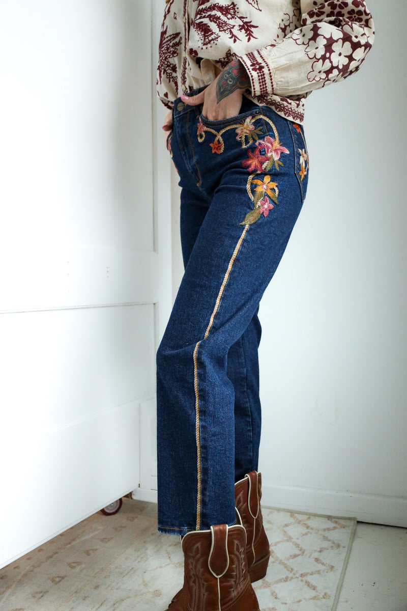 Person wearing blue jeans with floral embroidery and brown boots in a minimal indoor setting.
