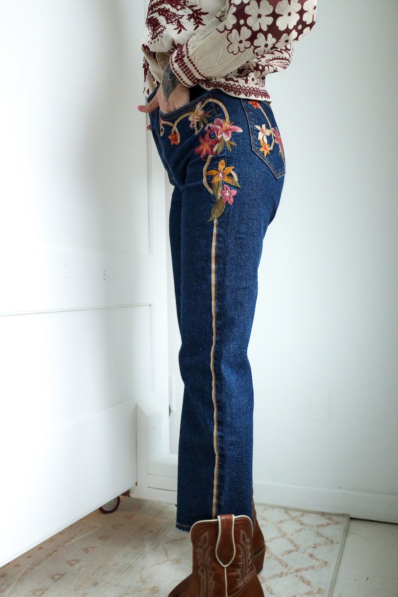 Person wearing blue jeans with floral embroidery and a patterned top, standing against a white wall.
