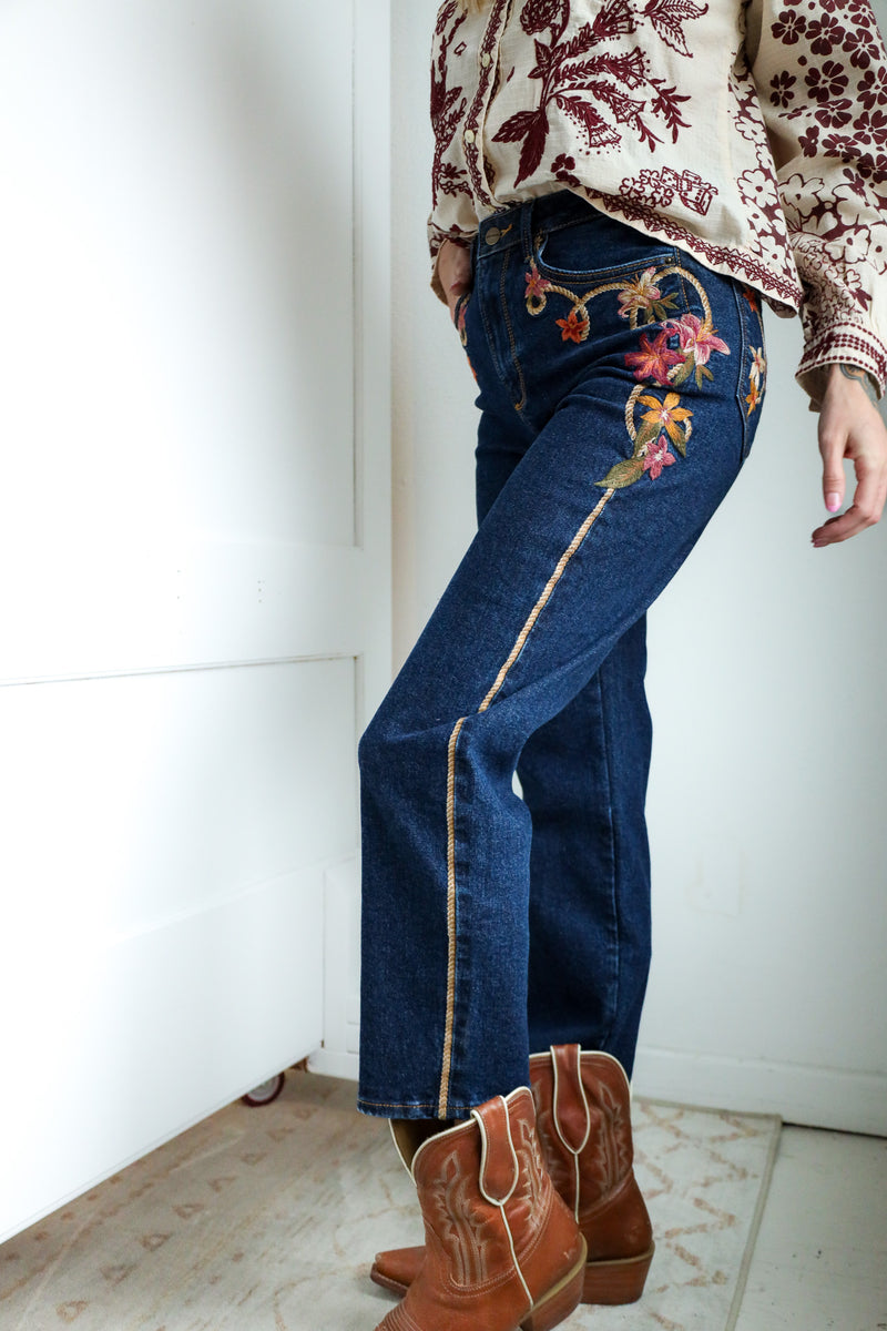 Person wearing blue jeans with floral embroidery and brown boots indoors.