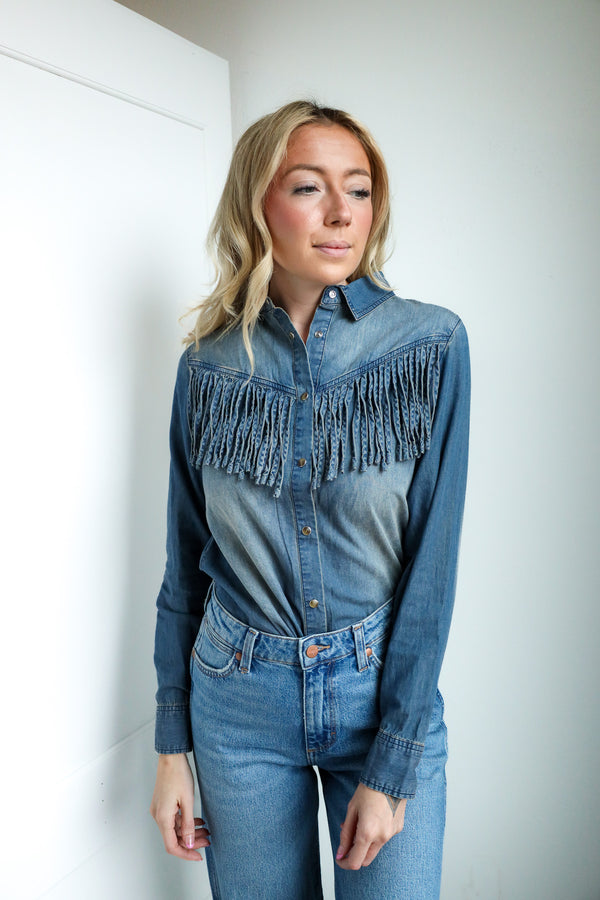 Woman wearing a denim outfit with a fringed shirt against a white wall