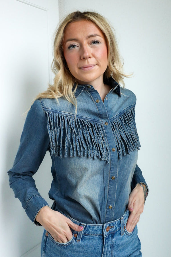 Woman wearing a blue denim shirt with fringe details and jeans against a white wall.