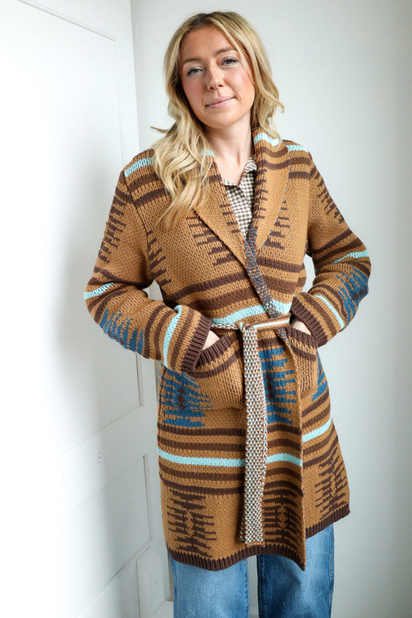 Woman wearing a patterned brown coat standing against a white wall.