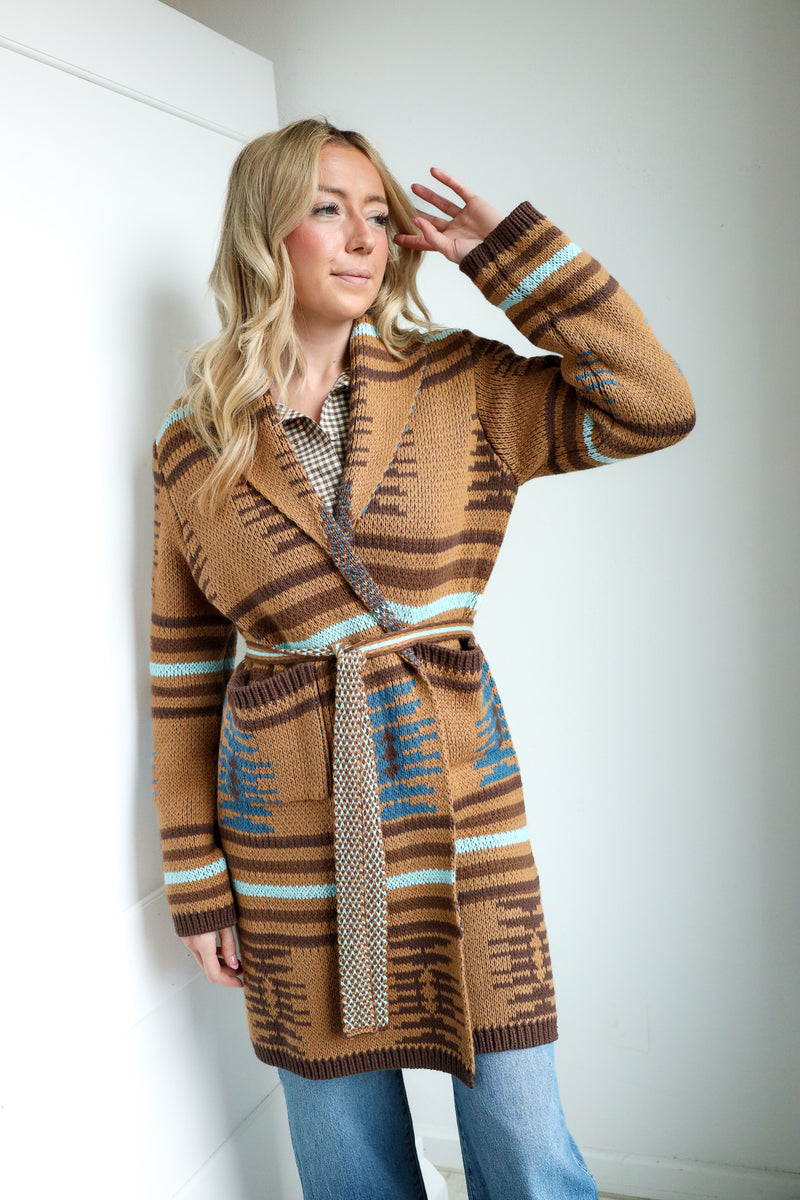 Woman wearing a patterned brown coat with a belt, standing against a white wall.