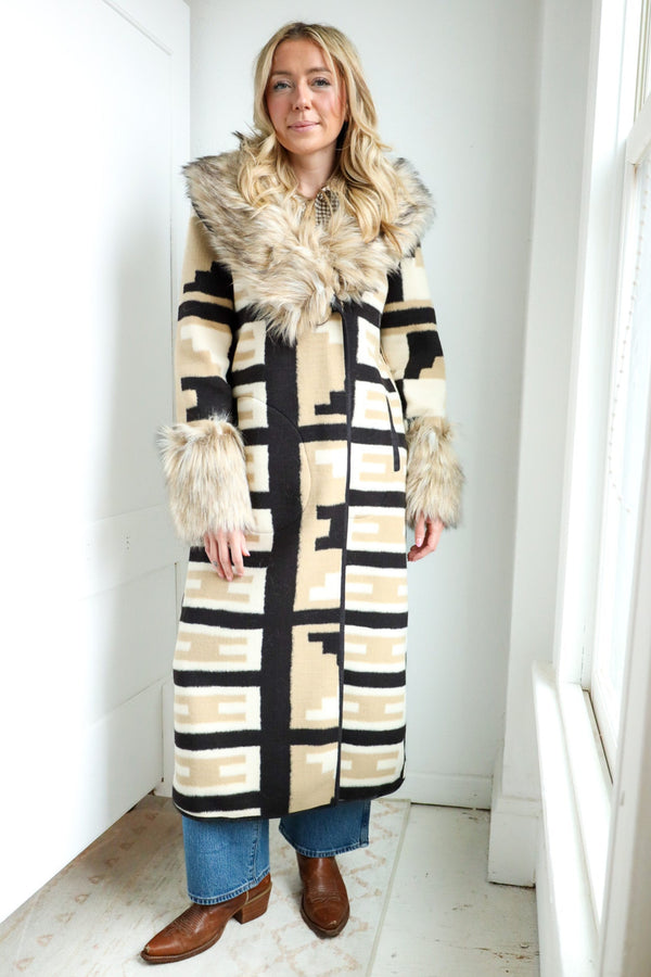 Person wearing a long, patterned coat with fur trim in a bright room.