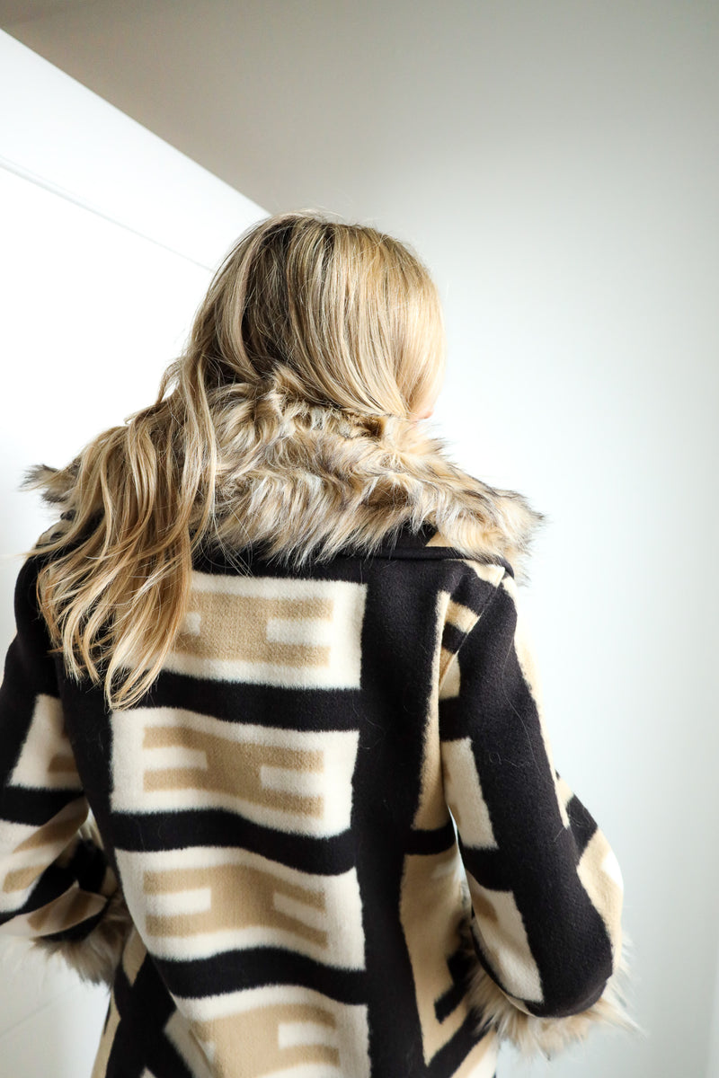 Person wearing a black and beige patterned coat with fur trim, standing against a plain background.