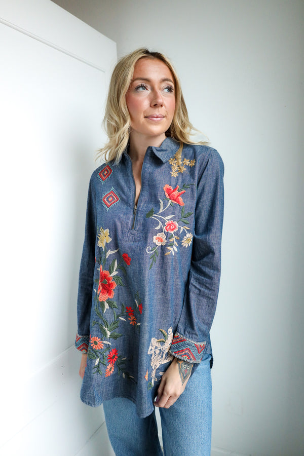 Woman wearing a blue denim shirt with floral embroidery against a white wall.