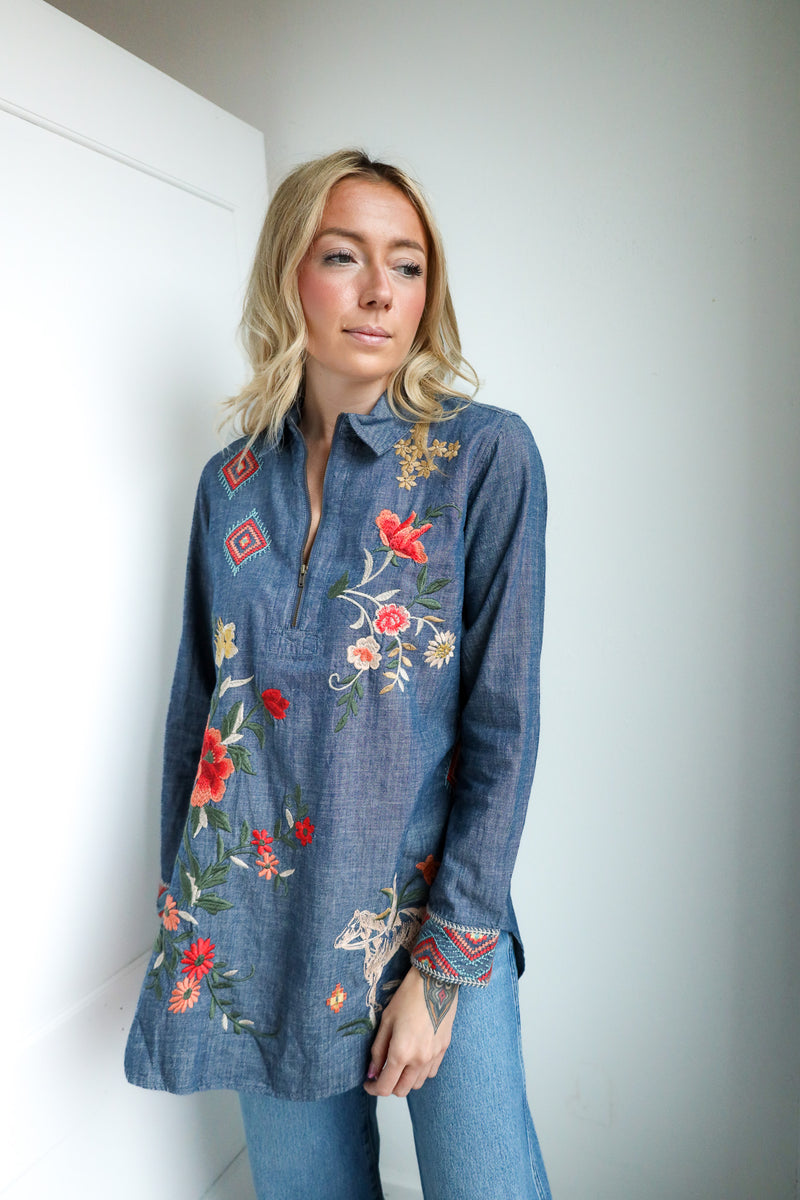Woman wearing a blue denim shirt with floral embroidery against a white wall.