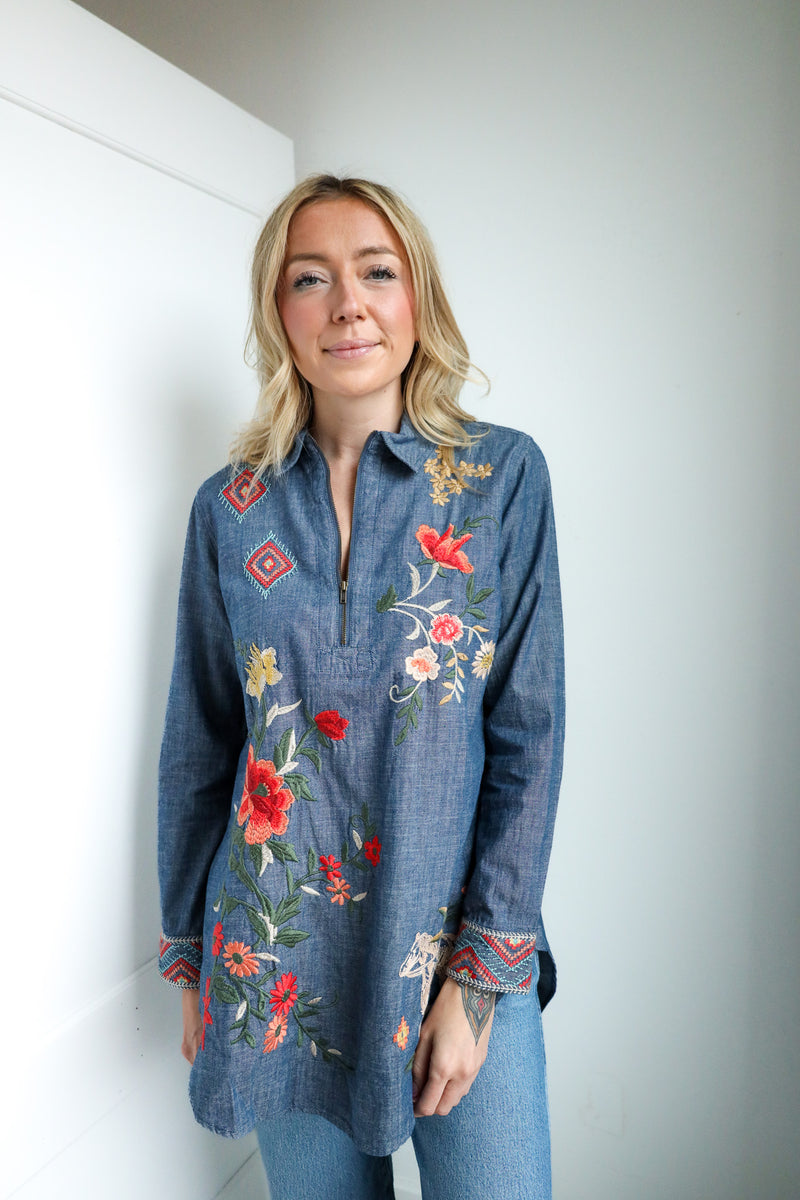 Woman wearing a blue denim shirt with floral embroidery against a white wall.