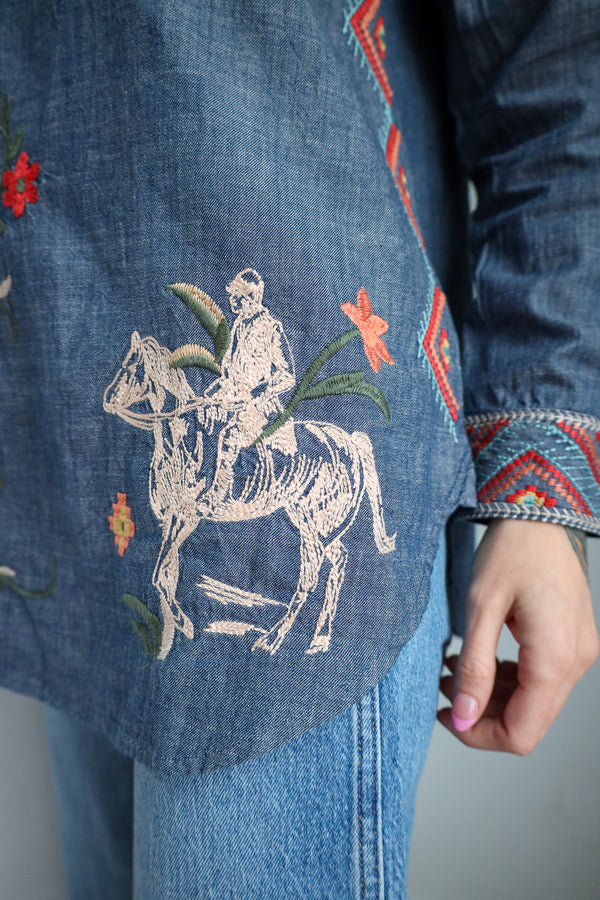 Blue denim jacket with embroidered design of a person on a horse and flowers.