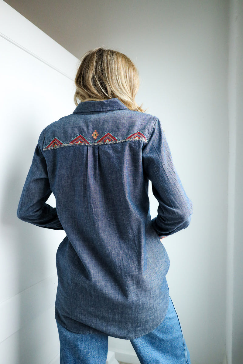 Person wearing a blue denim shirt with embroidered patterns, standing against a plain wall.