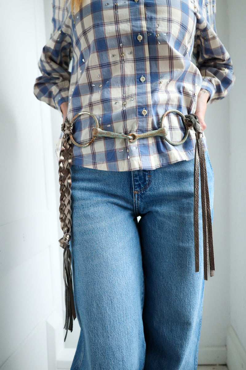 Person wearing a plaid shirt and blue jeans with a horse bit belt.