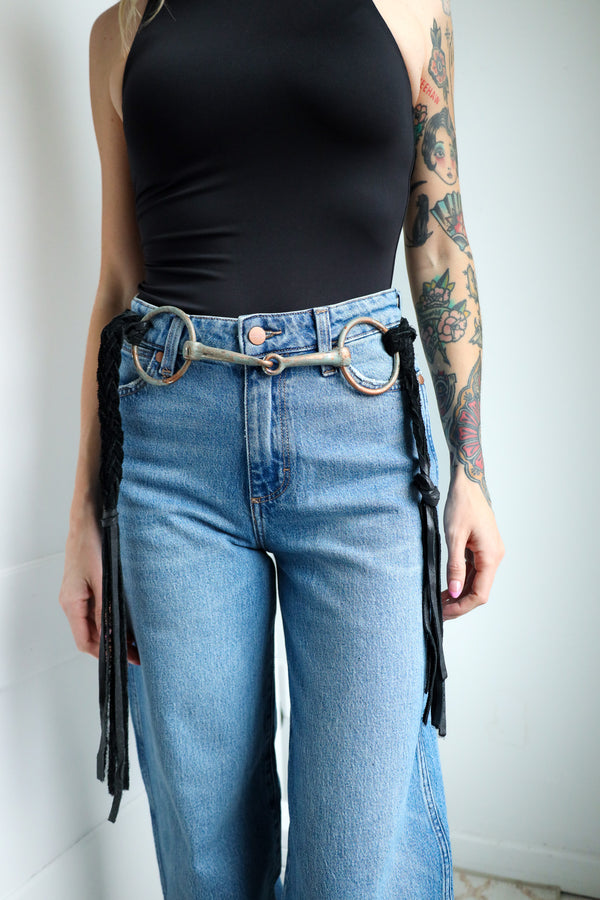Person wearing blue jeans with a decorative belt featuring fringe and a metallic buckle.