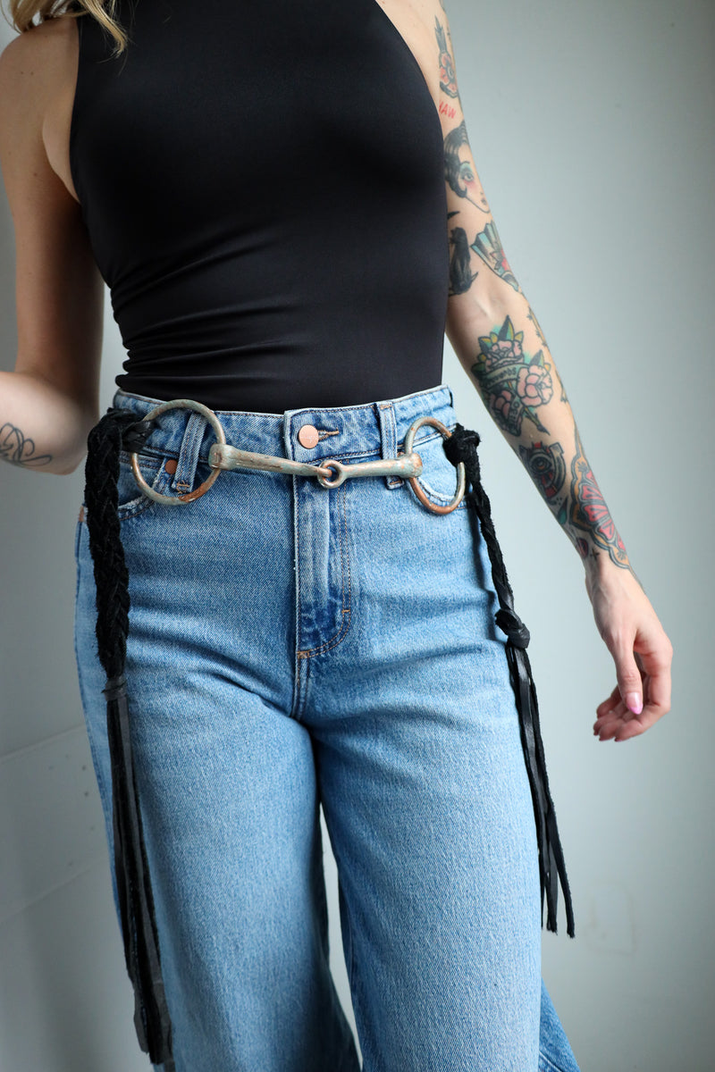 Person wearing blue jeans with a decorative belt and black fringes on a gray background