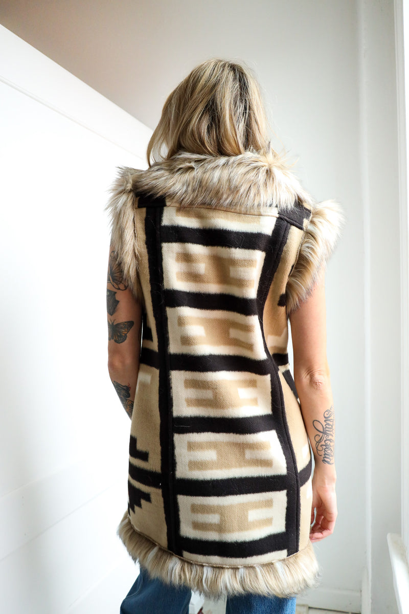 Person wearing a patterned vest with fur trim, standing against a white wall.