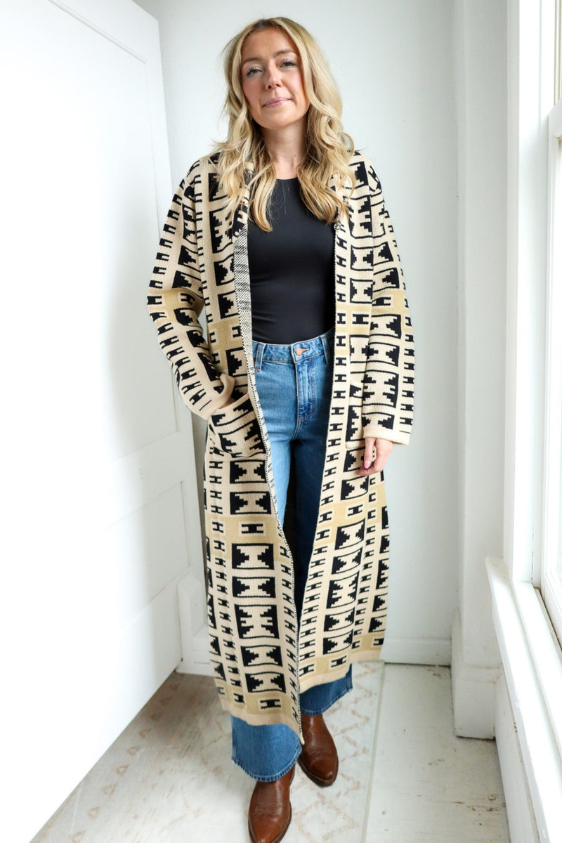 Woman wearing a long, patterned cardigan in a bright room.