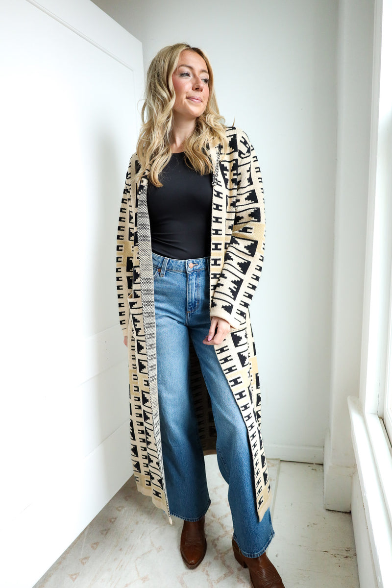 Woman wearing a long, patterned coat over a black top and blue jeans in a bright room.