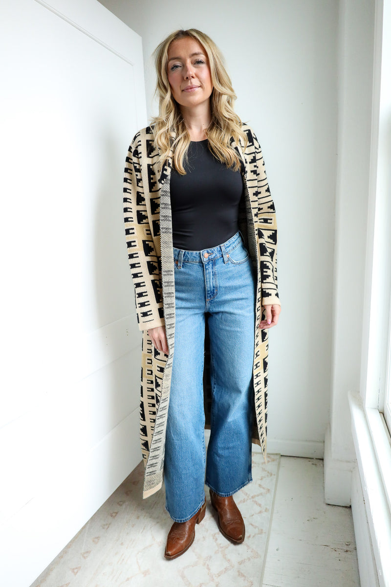 Woman wearing a long, patterned cardigan over a black top and blue jeans indoors.