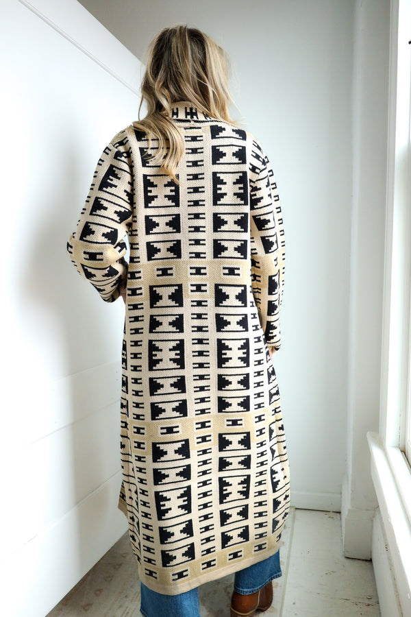 Person wearing a long, patterned coat standing in a room with white walls and a window.