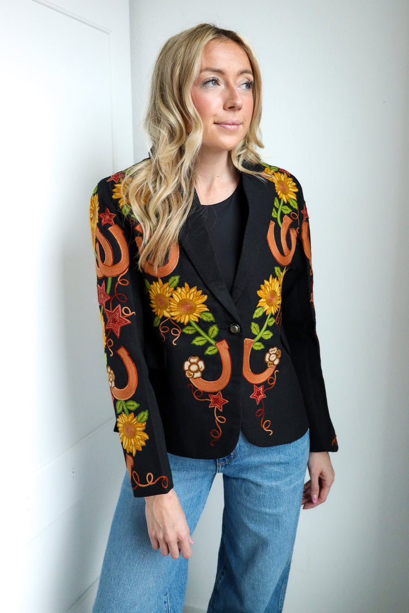 Woman wearing a black blazer with colorful floral and horseshoe patterns against a white wall.