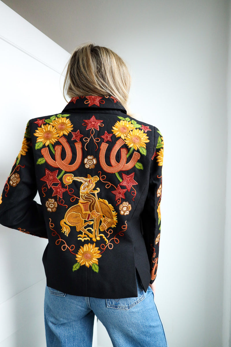 Person wearing a black jacket with colorful floral and horse embroidery against a white wall.