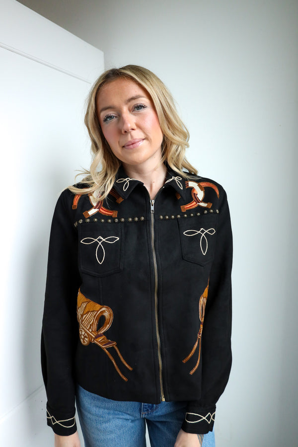Woman wearing a black jacket with orange embroidery against a white wall