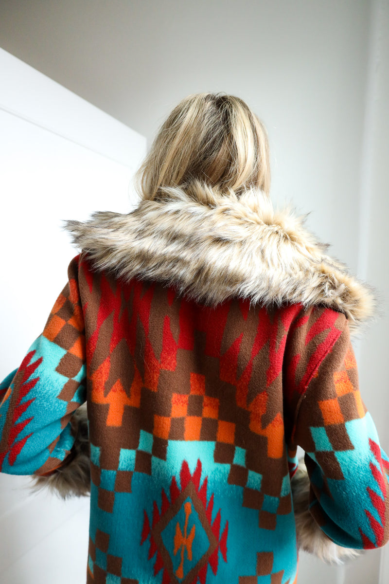 Person wearing a colorful patterned coat with fur trim on a plain background