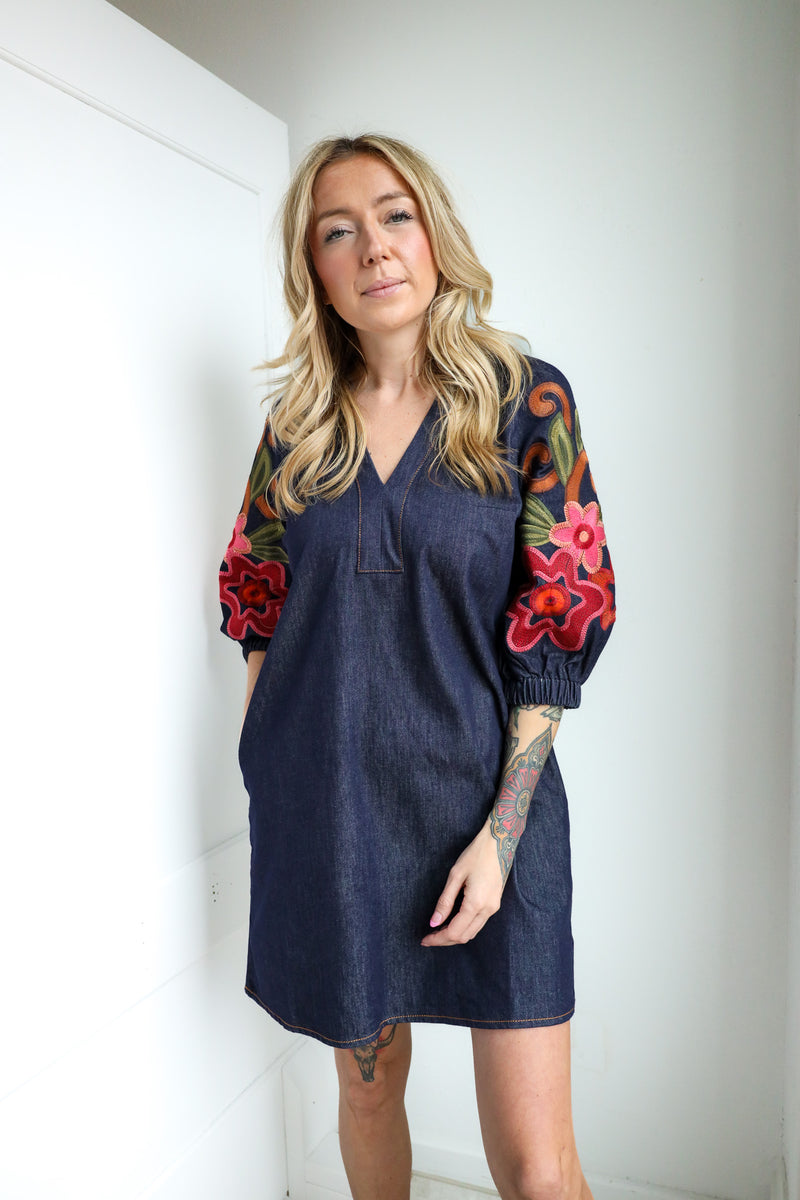 Woman wearing a denim dress with colorful floral sleeves against a white wall.