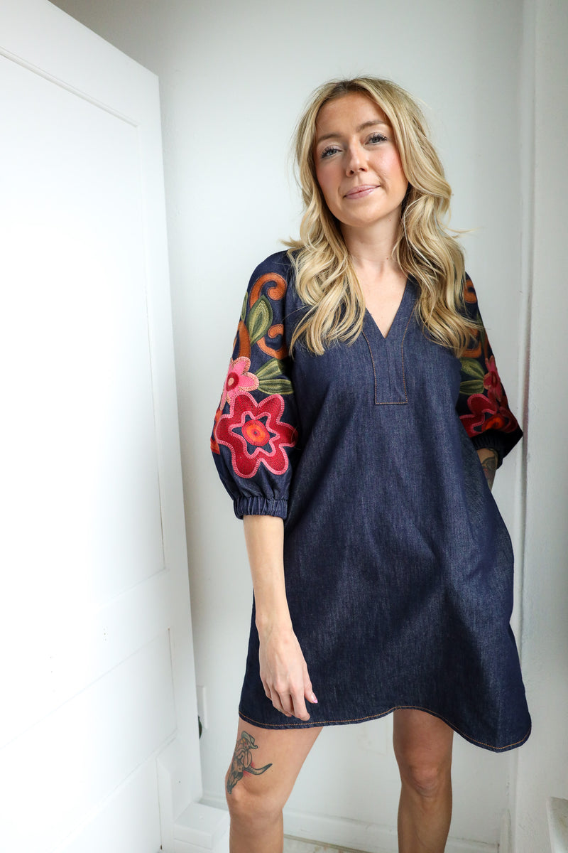 Woman wearing a denim dress with colorful floral sleeves against a white wall.