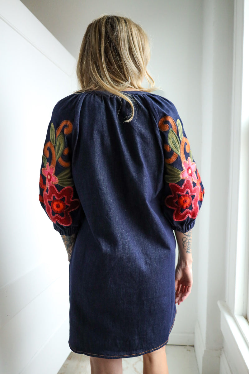 Person wearing a navy dress with colorful floral embroidery in a bright room.