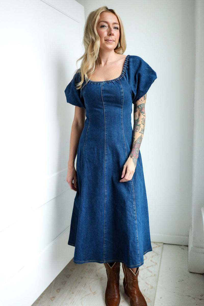 Woman wearing a blue denim dress standing against a white wall.