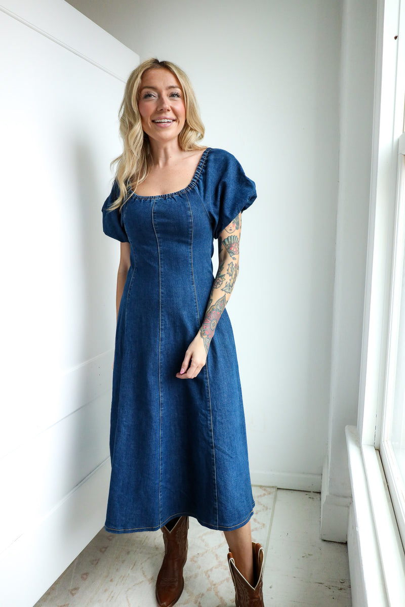 Woman wearing a blue denim dress standing against a white wall.