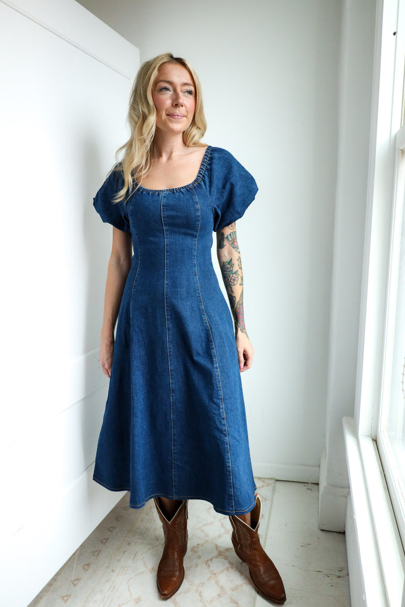 Woman wearing a blue dress standing in a room with white walls and a window.