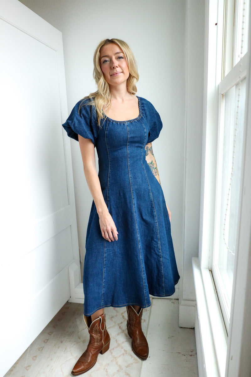 Woman wearing a blue denim dress standing in a bright room with white walls and a window.