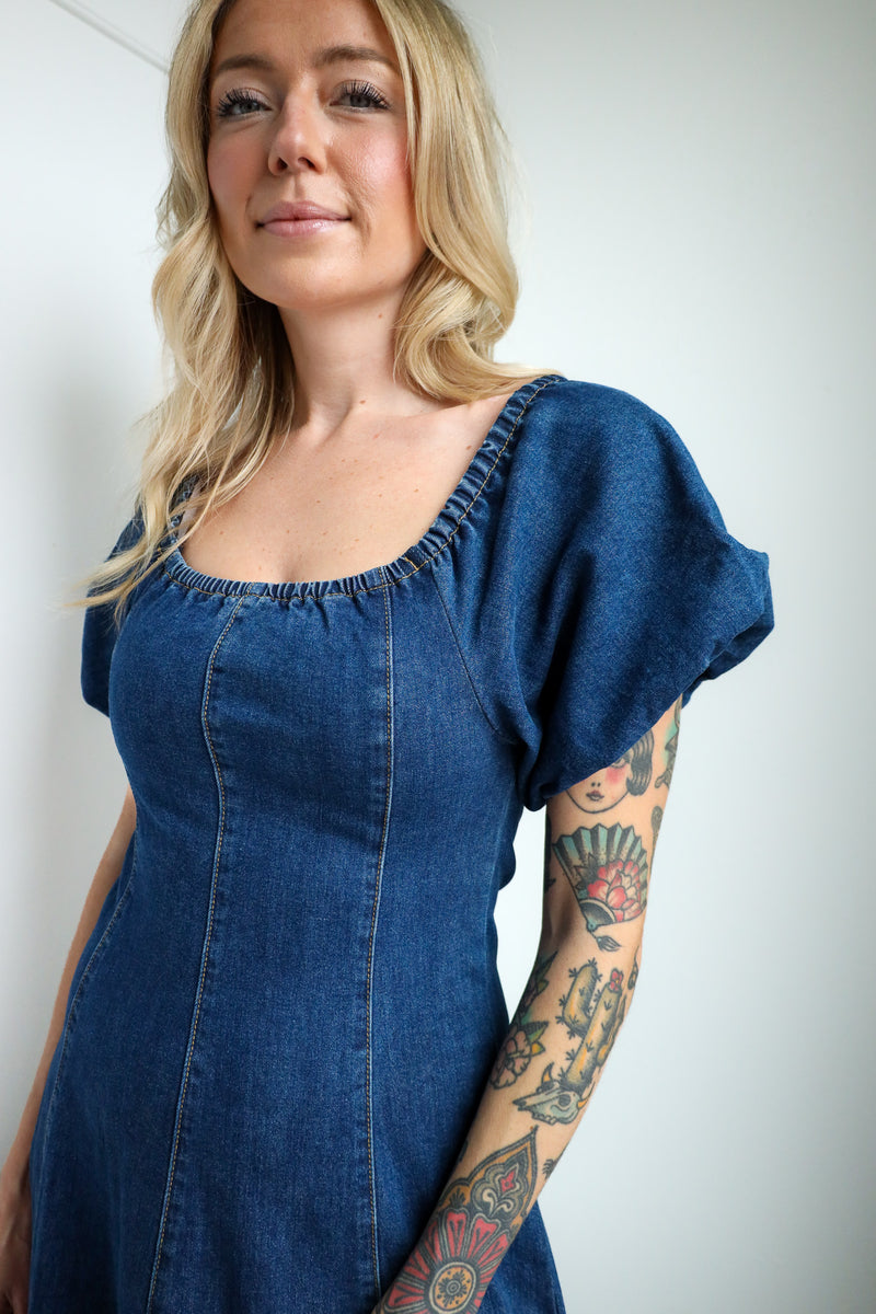 Woman wearing a blue denim dress with tattoos on her arms against a plain background