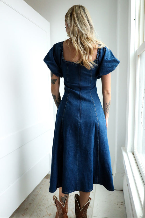 Woman wearing a blue dress and cowboy boots standing in a bright room with large windows.