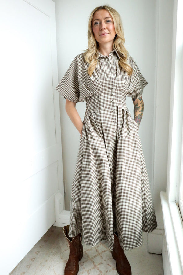 Woman wearing a checkered dress standing in a bright room with white walls and a window.