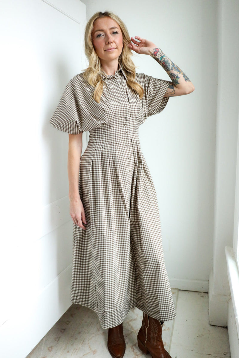 Woman wearing a checkered dress standing in a room with white walls and a wooden floor.