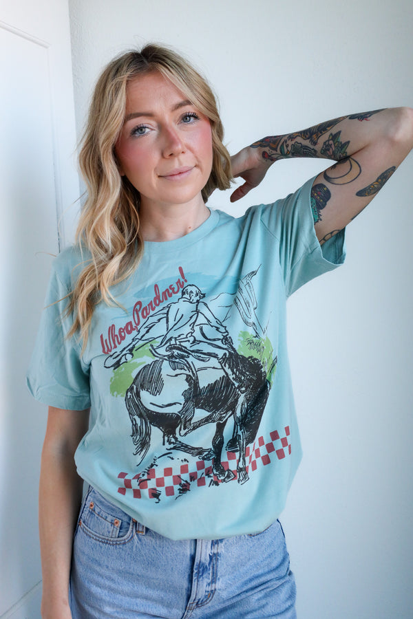 Woman wearing a light blue t-shirt with a graphic design of a person riding a horse, standing against a white wall.