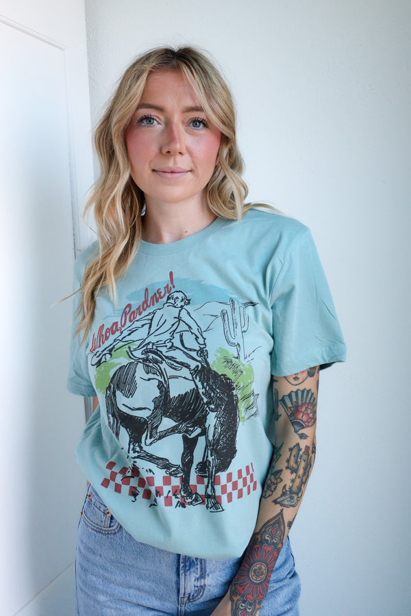 Woman wearing a light blue t-shirt with a graphic design of a person riding a horse, standing against a white wall.