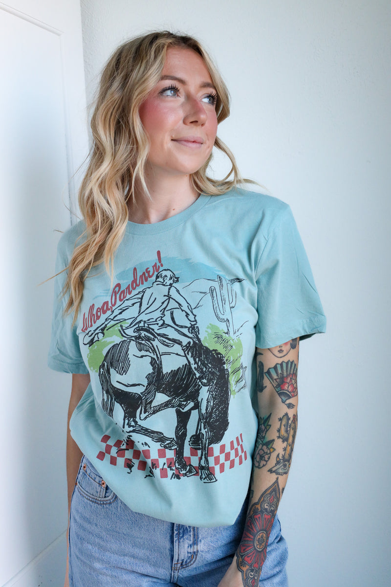 Woman wearing a light blue t-shirt with a graphic design of a person on a horse, standing against a white wall.