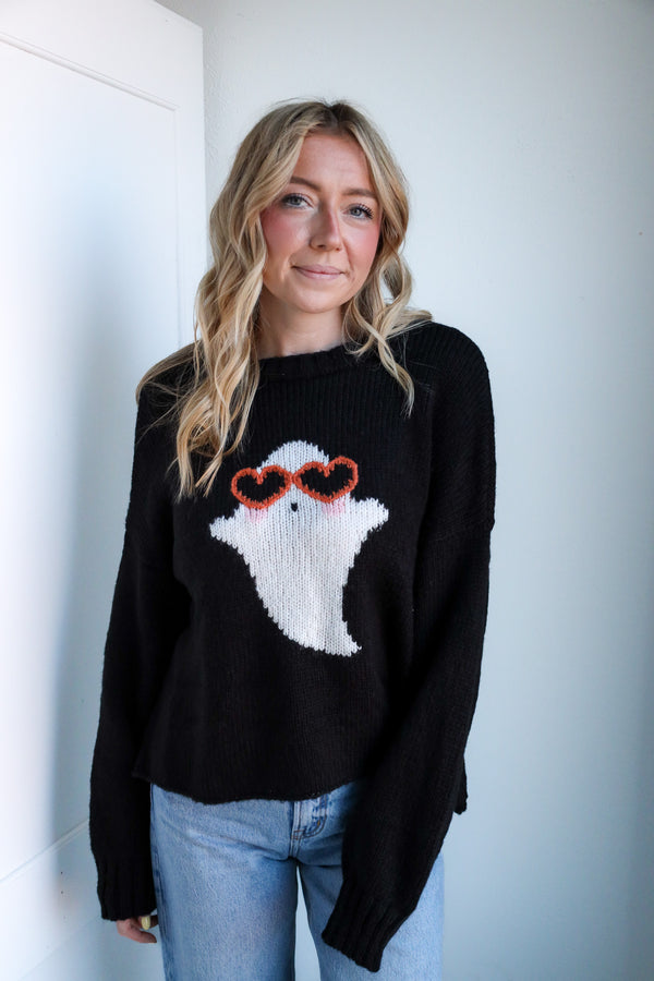 Woman wearing a black sweater with a ghost design, standing against a white wall.