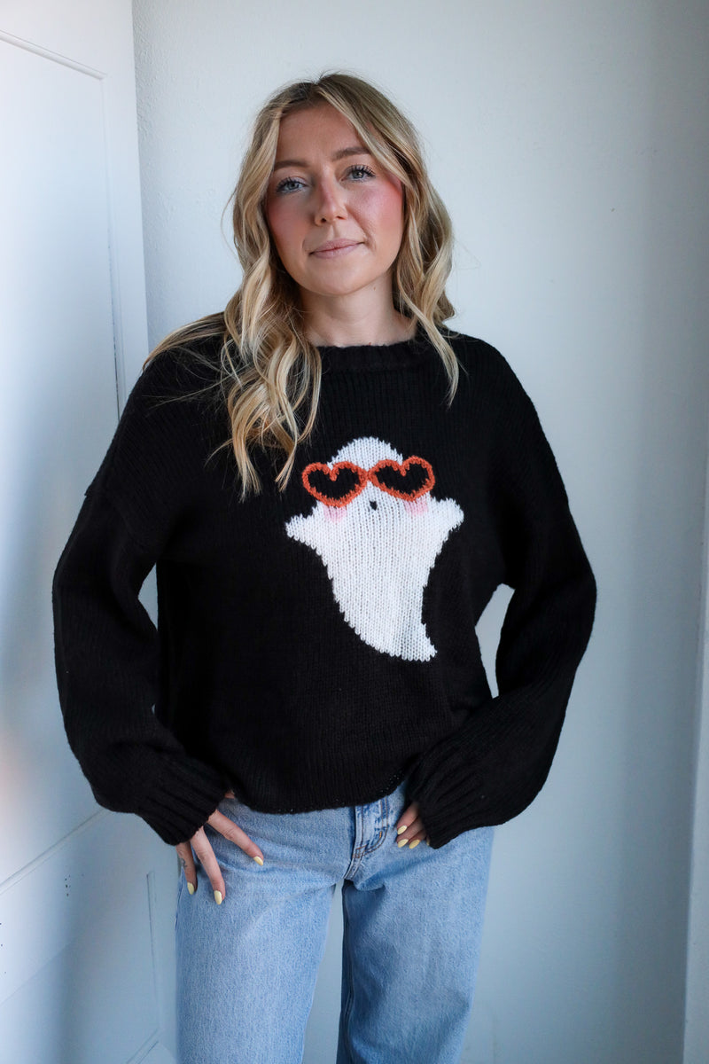 Person wearing a black sweater with a ghost design and sunglasses, standing against a white wall.