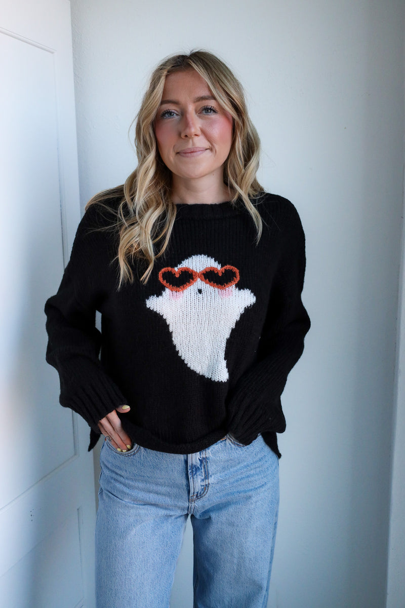 Person wearing a black sweater with a ghost design, standing against a white wall.