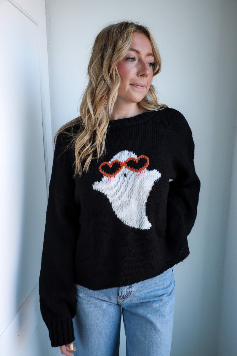 Person wearing a black sweater with a ghost design, standing against a white wall.