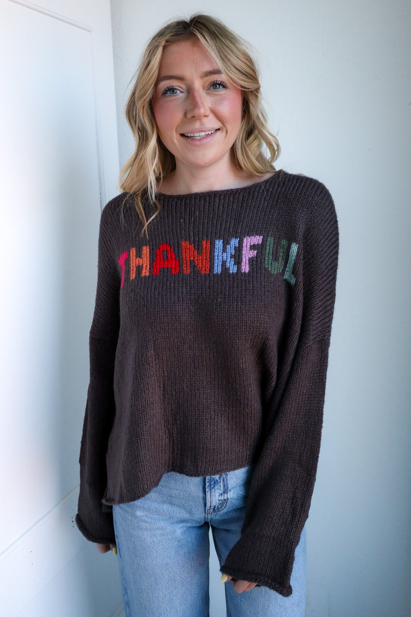 Woman wearing a brown sweater with 'THANKFUL' in colorful letters against a white wall.